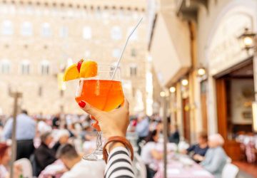 a woman lifting an aperol spritz into the sky on a patio in a city in italy